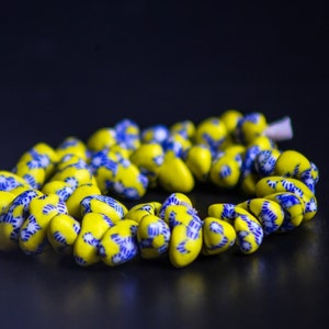 Op de afbeelding: Een ketting van gele kralen met blauwe bloempatronen. De druppelvormige kralen zijn aan elkaar geregen. De donkere achtergrond accentueert de heldere kleuren van de ketting, perfect voor een cadeau.
