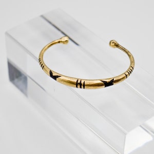 Puede incluir: Un brazalete de color dorado con diseños geométricos negros. El brazalete abierto se exhibe sobre un soporte de acrílico transparente. El brazalete presenta una serie de líneas verticales y formas de flecha.