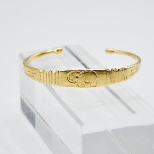 Peut inclure: Un bracelet manchette doré avec un motif d'éléphant en relief. Le bracelet a une section centrale rectangulaire avec des lignes verticales et des motifs floraux sur les côtés. Le bracelet est présenté sur une surface claire et inclinée.