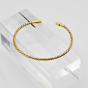 Peut inclure: Un bracelet jonc ouvert de couleur or avec un motif torsadé. Le bracelet est présenté sur un présentoir rectangulaire transparent sur fond blanc. Les extrémités du bracelet sont recouvertes d'or lisse. Bijoux.