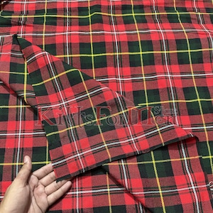 Pode incluir: Tecido tartan vermelho, verde e amarelo com uma faixa branca. O tecido está dobrado ao meio e uma mão está segurando o canto do tecido.