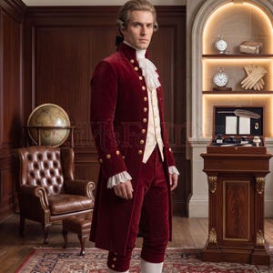Side-angle view of a burgundy velvet Georgian coat with a structured standing collar, gold button detailing, and white lace jabot, paired with a cream waistcoat and matching breeches. Photographed in a wood-paneled study with antique curios, this handmade colonial-style coat suits historical reenactors, Bridgerton fans, and 18th century costume collectors.