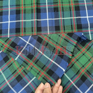 以下が含まれることがあります： 緑、青、黒、赤、白のチェック柄の生地のクローズアップ。生地は、交差する線を持つ伝統的なタータンパターンです。「Kilts For Men Store」の文字が生地に表示されています。