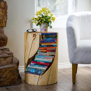 May include: A rustic, cylindrical wooden side table with a carved-out section for book storage. The table holds a stack of books, a potted plant with yellow flowers, and a pair of eyeglasses. The table is next to a white armchair.