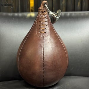 以下が含まれることがあります： 中央に縫い目のある、茶色の革製ティアドロップ型のスピードバッグ。上部にループと、吊り下げ用の金属製スイベルが付いています。ボクシングや武道のトレーニング用に設計されています。