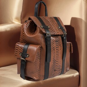K&ouml;nnte beinhalten: Brauner Rucksack aus Kunstschlangenleder mit schwarzen Riemen und Akzenten. Er verf&uuml;gt &uuml;ber einen oberen Griff, zwei Vordertaschen und verstellbare Tr&auml;ger. Die Tasche hat einen Klappenverschluss mit Schnallen und ein strukturiertes Muster.