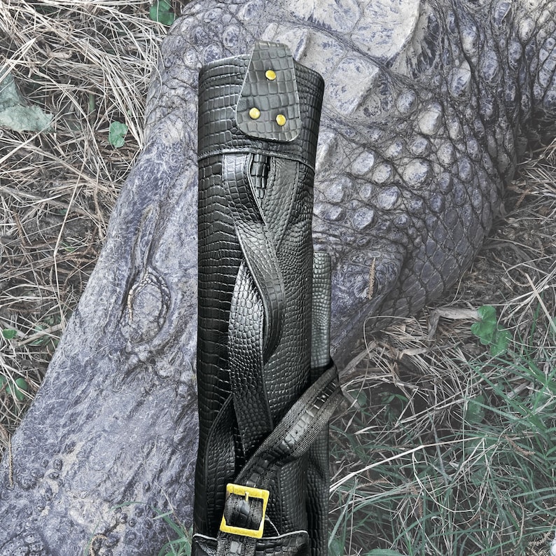 Pu&ograve; includere: Borsa da golf in pelle nera con motivo coccodrilo e dettagli dorati. La borsa ha una chiusura a fibbia e una cerniera.