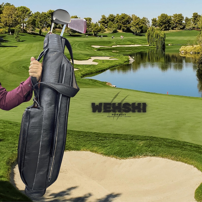 Pu&ograve; includere: Una sacca da golf in pelle nera con chiusura a zip. La sacca &egrave; tenuta da una persona su un campo da golf con uno stagno e erba verde sullo sfondo. La sacca ha il testo "WEKSHI PURSUIT REBORNCONTEMPORARY" su di essa.