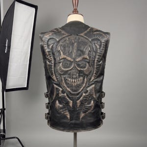 Könnte beinhalten: Eine schwarze Lederweste mit einem großen, geprägten Totenkopf-Design auf der Rückseite mit abgenutzten braunen Highlights. Das Kleidungsstück enthält dekorative Steppmuster um den Totenkopf und drei verstellbare Seitengurte mit Metallschnallen.