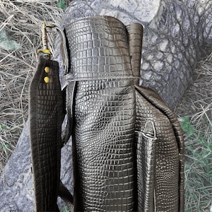 Pu&ograve; includere: Una borsa da golf in pelle nera con un motivo a coccodrillo. La borsa ha una tracolla e una tasca con cerniera.