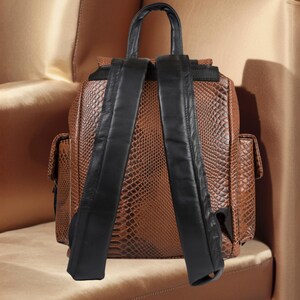 K&ouml;nnte beinhalten: Brauner Rucksack aus Kunstleder mit Schlangenmuster und schwarzen Lederriemen und Griff. Der Rucksack hat zwei Seitentaschen und einen Klappenverschluss. Die Tasche steht auf einer goldfarbenen Oberfl&auml;che.