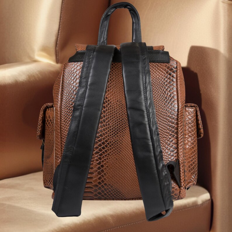 K&ouml;nnte beinhalten: Brauner Rucksack aus Kunstleder mit Schlangenmuster und schwarzen Lederriemen und Griff. Der Rucksack hat zwei Seitentaschen und einen Klappenverschluss. Die Tasche steht auf einer goldfarbenen Oberfl&auml;che.