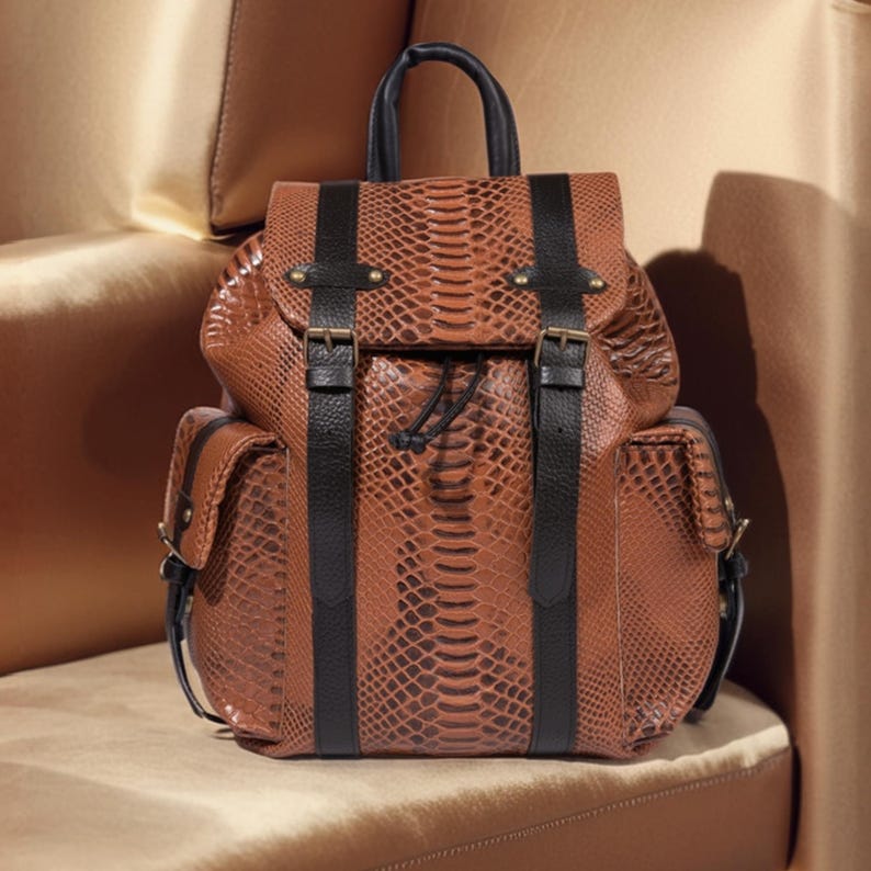 K&ouml;nnte beinhalten: Ein brauner Lederrucksack mit Schlangenmuster. Der Rucksack hat schwarze Lederriemen, einen oberen Griff und zwei Seitentaschen. Die Tasche wird mit Schnallen und einem Kordelzugverschluss gesichert. Die Tasche steht auf einer beigen Oberfl&auml;che.