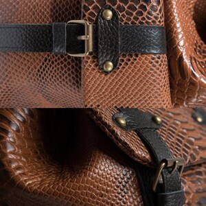 K&ouml;nnte beinhalten: Nahaufnahme einer braunen Ledertasche mit Schlangenmuster. Die Tasche hat schwarze Lederriemen und goldfarbene Metallbeschl&auml;ge, einschlie&szlig;lich Schnallen und Nieten. Die Textur des Leders ist sichtbar.