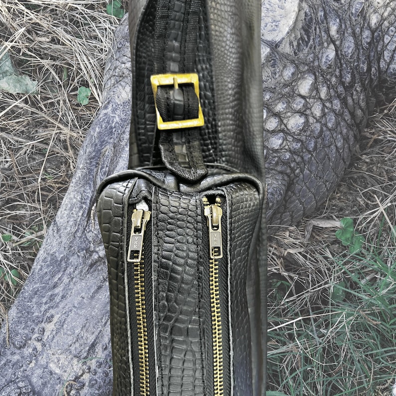 Pu&ograve; includere: Una borsa a tracolla in pelle nera con una fibbia dorata e due cerniere dorate. La borsa ha un motivo goffrato a coccodrillo.