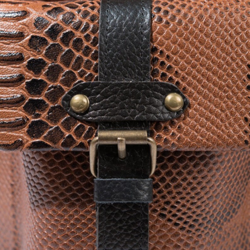 K&ouml;nnte beinhalten: Nahaufnahme einer braunen und schwarzen Ledertasche mit Schlangenmuster. Die Tasche hat einen schwarzen Lederriemen mit einer goldfarbenen Schnalle und Messingnieten. Das Leder hat ein strukturiertes Aussehen.