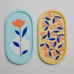 Puede incluir: Dos bandejas ovaladas para servir con diseños diferentes. Una bandeja es azul claro con fondo blanco y una flor naranja estilizada con hojas azules. La otra bandeja es amarilla con fondo blanco y un patrón de hojas azules y bayas naranjas.