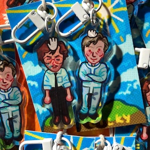 May include: Two colorful cartoon character keychains with white key rings. The characters are wearing blue and white outfits and have brown hair. The background is a blue sky with white clouds and yellow sun rays.