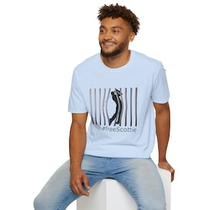 Puede incluir: Una camiseta azul claro con una silueta negra de un golfista balanceando un palo, rompiendo un conjunto de barras verticales. El texto '#freeScottie' está impreso debajo de la imagen.