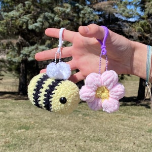 May include: Two crocheted keychains, one shaped like a bee with black and yellow stripes and a white fuzzy head, and the other shaped like a pink flower with a yellow center.