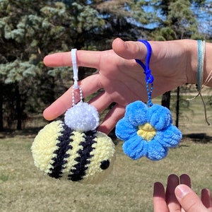 May include: Two crocheted keychains, one shaped like a bee with yellow and black stripes and a white pom-pom, and the other shaped like a blue flower with a yellow center.