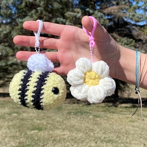 May include: A crocheted bee and daisy keychain set. The bee is yellow and black with a white fuzzy head. The daisy is white with a yellow center. Both are attached to pink and white plastic keychains.