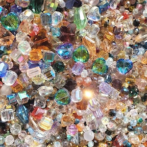 May include: A close-up view of a collection of colorful, faceted beads. The beads are in various shapes, including hearts, squares, and rounds. Colors include green, blue, orange, purple, and clear. The beads are scattered, creating a sparkling, textured surface.
