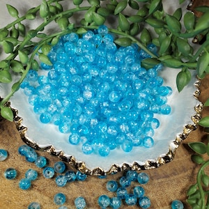 Pode incluir: Uma cole&ccedil;&atilde;o de contas de vidro azul claro com efeito craquelado. As contas redondas est&atilde;o espalhadas numa tigela branca e dourada, com folhagem verde no fundo. Estas contas s&atilde;o adequadas para fazer joias e artesanato.