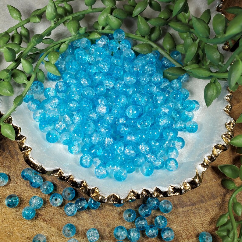 Pode incluir: Uma cole&ccedil;&atilde;o de contas de vidro azul claro com efeito craquelado. As contas redondas est&atilde;o espalhadas numa tigela branca e dourada, com folhagem verde no fundo. Estas contas s&atilde;o adequadas para fazer joias e artesanato.