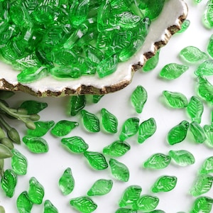 May include: A collection of small, translucent emerald green glass beads shaped like leaves. The beads feature a detailed vein pattern on their surface and are scattered across the frame, with many contained within a white dish with gold trim.