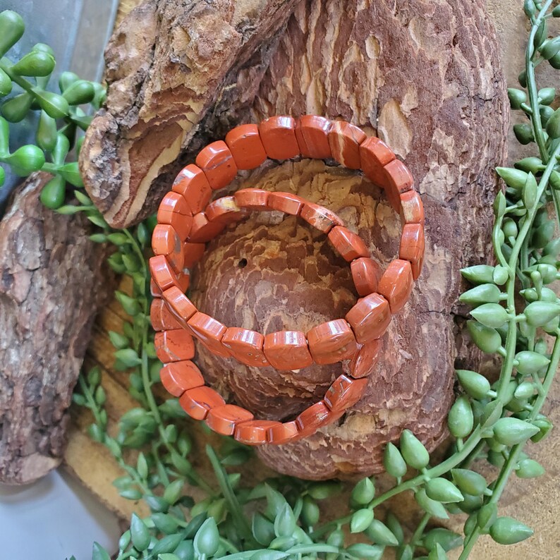K&ouml;nnte beinhalten: Zwei orange-rote Steinarmb&auml;nder mit rechteckigen Perlen werden auf einer Holzoberfl&auml;che pr&auml;sentiert, umgeben von gr&uuml;nen Pflanzenakzenten. Die Armb&auml;nder sind gestapelt und zeigen ihre erdigen T&ouml;ne und ihre nat&uuml;rliche Textur. Ein einzigartiges Accessoire.