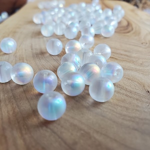 May include: A collection of round, translucent white beads with an iridescent finish that reflects subtle blue and pink hues. Each bead features a smooth, spherical surface and a central hole, scattered across the frame in a loose arrangement.