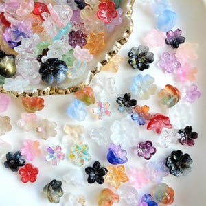 May include: A collection of small, translucent flower-shaped beads in a variety of colors including clear, pink, blue, orange, purple, and black. Some beads feature iridescent finishes, glitter flecks, or gradient color transitions.