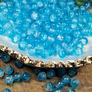 Pode incluir: Uma cole&ccedil;&atilde;o de contas de vidro azul claro com efeito craquelado. As contas s&atilde;o redondas e transparentes, com uma apar&ecirc;ncia texturizada e fraturada. Elas s&atilde;o exibidas em uma tigela decorativa com borda dourada.