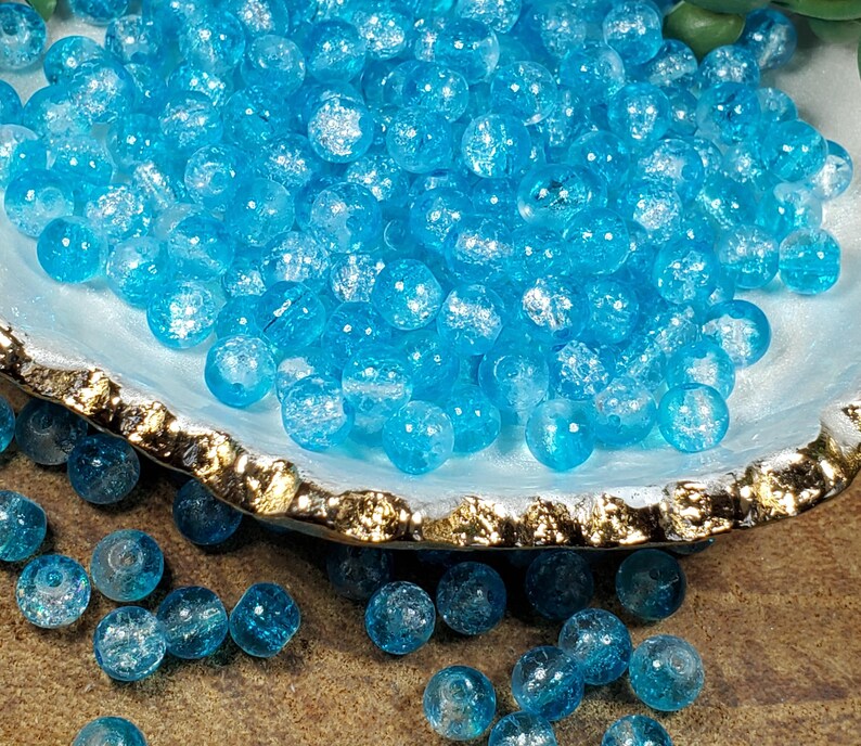 Pode incluir: Uma cole&ccedil;&atilde;o de contas de vidro azul claro com efeito craquelado. As contas s&atilde;o redondas e transparentes, com uma apar&ecirc;ncia texturizada e fraturada. Elas s&atilde;o exibidas em uma tigela decorativa com borda dourada.