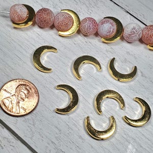 Puede incluir: Encantos dorados en forma de media luna y cuentas rosas y blancas estampadas. Se incluye una moneda para comparar el tama&ntilde;o. Los encantos est&aacute;n sobre una superficie de madera clara, lo que sugiere suministros para hacer joyas.