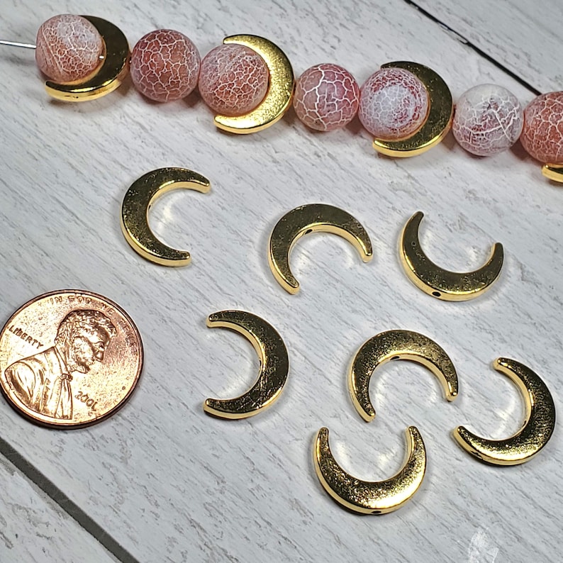 Puede incluir: Encantos dorados en forma de media luna y cuentas rosas y blancas estampadas. Se incluye una moneda para comparar el tama&ntilde;o. Los encantos est&aacute;n sobre una superficie de madera clara, lo que sugiere suministros para hacer joyas.
