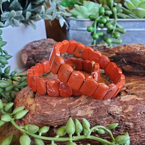 K&ouml;nnte beinhalten: Zwei orange-rote Steinarmb&auml;nder mit rechteckigen Perlen. Die Armb&auml;nder sind auf einem Holzst&uuml;ck gestapelt, umgeben von Gr&uuml;n. Die Armb&auml;nder sind ein lebendiges Accessoire.