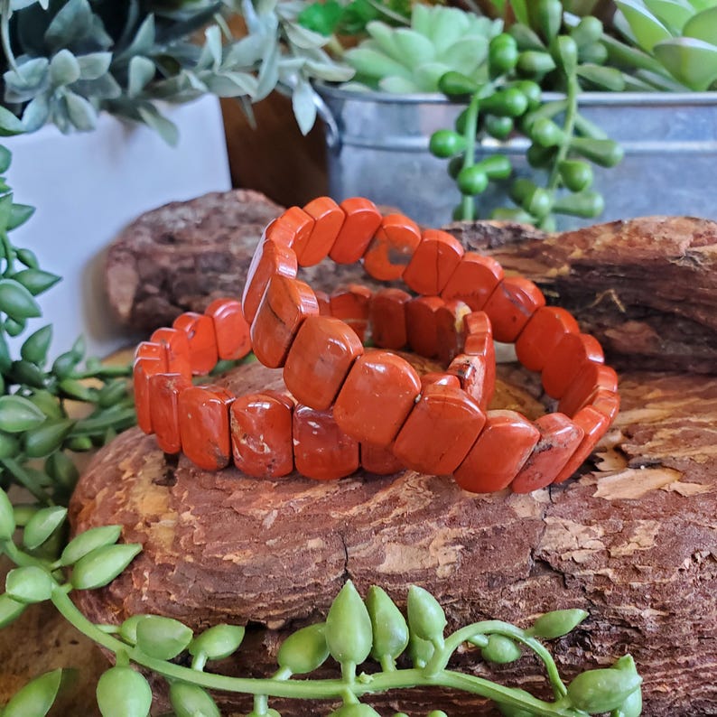 K&ouml;nnte beinhalten: Zwei orange-rote Steinarmb&auml;nder mit rechteckigen Perlen. Die Armb&auml;nder sind auf einem Holzst&uuml;ck gestapelt, umgeben von Gr&uuml;n. Die Armb&auml;nder sind ein lebendiges Accessoire.