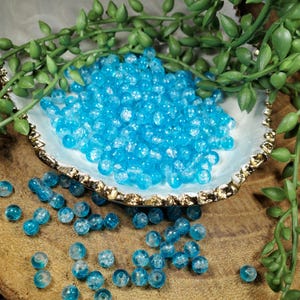 Pode incluir: Um conjunto de contas de vidro azul claro com efeito craquelado. As contas est&atilde;o espalhadas sobre uma superf&iacute;cie de madeira e num prato decorativo com borda dourada e branca, com folhagem verde no fundo. Estas contas s&atilde;o adequadas para a cria&ccedil;&atilde;o de joias e artesanato.