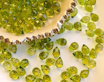 Puede incluir: Una colección de pequeñas cuentas de vidrio de color verde lima facetadas con forma de lágrima. Muchas cuentas están agrupadas dentro de un plato de cerámica blanco roto, curvo y texturizado con borde dorado, otras dispersas.