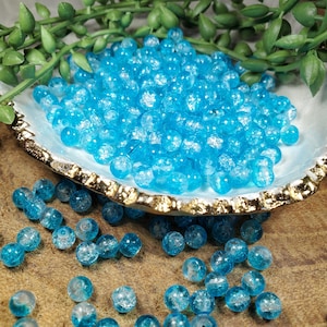 Pode incluir: Uma cole&ccedil;&atilde;o de contas de vidro azul brilhante com efeito craquelado. As contas s&atilde;o redondas e est&atilde;o espalhadas numa tigela decorativa em forma de concha com uma borda dourada. A folhagem verde &eacute; vis&iacute;vel no fundo. As contas t&ecirc;m cerca de 0,8 cm de di&acirc;metro.