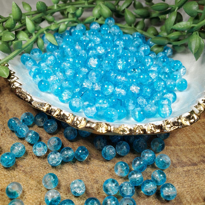 Pode incluir: Uma cole&ccedil;&atilde;o de contas de vidro azul brilhante com efeito craquelado. As contas s&atilde;o redondas e est&atilde;o espalhadas numa tigela decorativa em forma de concha com uma borda dourada. A folhagem verde &eacute; vis&iacute;vel no fundo. As contas t&ecirc;m cerca de 0,8 cm de di&acirc;metro.