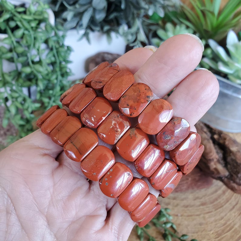 K&ouml;nnte beinhalten: Drei Reihen rotbrauner Jaspis-Steinarmb&auml;nder. Die rechteckigen Steine sind poliert und weisen Farb- und Musterunterschiede auf. Die Armb&auml;nder werden in einer Hand gehalten, mit einem unscharfen Hintergrund aus Gr&uuml;n.