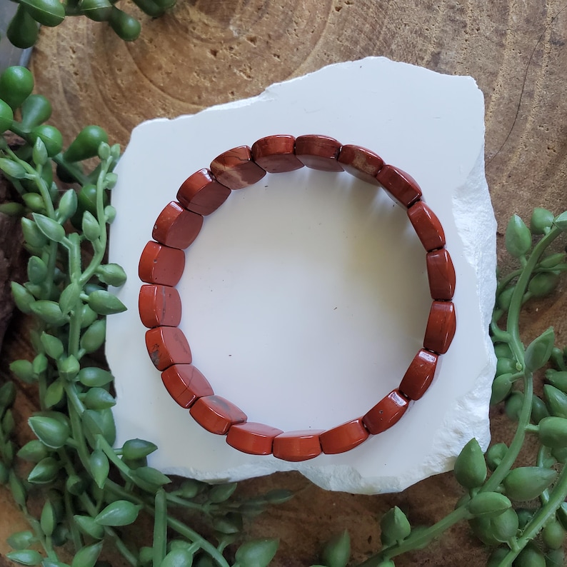 K&ouml;nnte beinhalten: Ein Armband aus rotem Jaspis mit rechteckigen Perlen. Das Armband wird auf einer wei&szlig;en, strukturierten Oberfl&auml;che pr&auml;sentiert, umgeben von gr&uuml;nen Pflanzensprossen und einem h&ouml;lzernen Hintergrund. Die Perlen haben eine polierte Oberfl&auml;che.