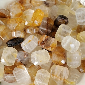 May include: A collection of polished, tumbled gemstones in shades of yellow, amber, and clear quartz. The stones vary in size and shape, with some showing internal inclusions and natural patterns. The gemstones are scattered, creating a textured surface.