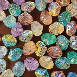 May include: A collection of iridescent glass beads shaped like seashells. The beads feature a translucent finish with a shimmering, multi-colored coating in shades of blue, green, purple, orange, and yellow, displaying a ribbed texture.