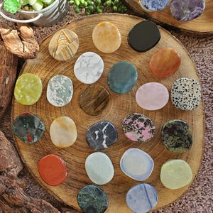 May include: A collection of polished gemstones in various colors and patterns, arranged on a wooden slice. The stones include shades of green, pink, brown, and blue, with unique veining and textures. The arrangement is on a brown surface with greenery in the background.