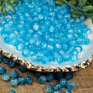 Pode incluir: Close-up de contas de vidro craquelado azul claro. As contas redondas t&ecirc;m um acabamento fosco e texturizado. S&atilde;o apresentadas numa concha com borda dourada. Ideais para fazer joias ou outros projetos de artesanato.