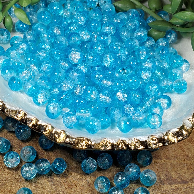 Pode incluir: Close-up de contas de vidro craquelado azul claro. As contas redondas t&ecirc;m um acabamento fosco e texturizado. S&atilde;o apresentadas numa concha com borda dourada. Ideais para fazer joias ou outros projetos de artesanato.